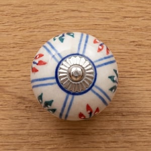 May include: A round, white ceramic cabinet knob with a silver metal base. The knob features a pattern of blue stripes, red and green floral accents. The knob is attached to a light brown wooden surface.