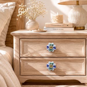 Könnte beinhalten: Ein helles Nachtschränkchen aus Holz mit zwei Schubladen, jede mit einem blumenförmigen Knopf. Die Knöpfe haben blaue und grüne Blütenblätter mit einem goldenen Zentrum. Eine Lampe, eine Kerze und Bücher stehen oben.