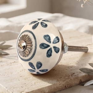 Peut inclure: Un bouton de meuble rond en céramique avec une base de couleur crème et des motifs floraux bleus peints à la main. Le centre présente une rosace décorative argentée avec une tige filetée métallique dépassant à l'arrière pour l'installation.