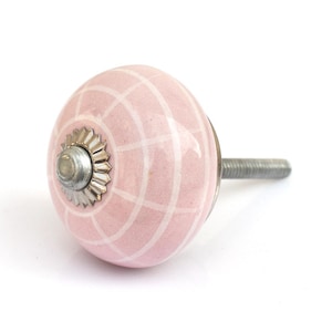May include: A round ceramic cabinet knob in a soft pink color featuring a white grid pattern. The knob has a silver-toned metal base with a scalloped edge and a threaded metal bolt extending from the back for installation on furniture or doors.