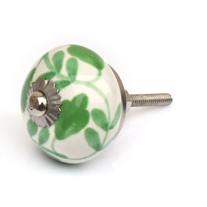 May include: A round ceramic cabinet knob featuring a white base with a hand-painted green leaf pattern. The center of the knob has a silver-toned metal decorative cap and a matching silver-toned threaded metal bolt extending from the back.
