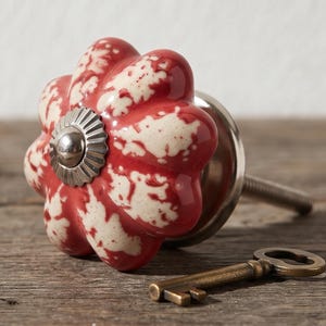 Puede incluir: Un pomo de cerámica para muebles con forma de flor, con un patrón moteado en rojo y blanco, una tapa central de metal plateado y un perno roscado, acompañado de una pequeña llave de latón de estilo antiguo situada cerca.