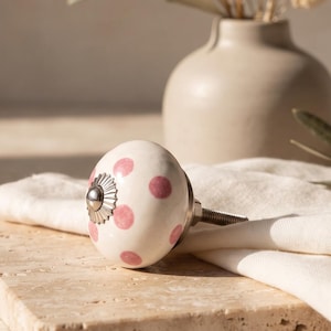 Op de afbeelding: Een ronde keramische meubelknop met een witte basis en een patroon van roze stippen. De knop heeft een zilverkleurig metalen accent in het midden en een metalen draadeind aan de achterzijde voor bevestiging op meubelstukken.