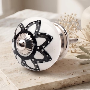 Può includere: Un pomello rotondo in ceramica bianca con un motivo floreale nero e piccoli punti bianchi lungo i petali. Il centro del pomello presenta un tappo metallico argentato con bordo scanalato e un bullone filettato sul retro.
