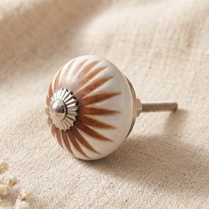 May include: A round ceramic cabinet knob featuring a white base with a brown sunburst pattern radiating from the center. The center of the knob is accented with a small, silver-toned metal cap with a fluted edge, and a threaded metal bolt extends from the back.