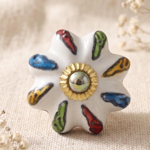 Puede incluir: Un pomo de cerámica blanca para muebles con forma de flor de ocho pétalos redondeados. Cada pétalo presenta un pequeño acento pintado alargado en rojo, azul, verde o amarillo. El centro tiene una placa de metal dorado festoneada.