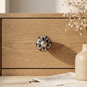 Könnte beinhalten: Ein runder Keramik-Schubladenknopf mit einem schwarz-weißen Blumenmuster und einem zentralen silberfarbenen Metallakzent, montiert an einer hellen Eichenholz-Schubladenfront, neben einer beigen Vase mit Trockenblumen und einem weißen Tuch.