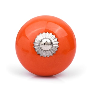 Può includere: Un pomello per mobili sferico di colore arancione con una finitura liscia e lucida. Il centro del pomello presenta una piastra metallica circolare color argento con un bordo smerlato e una piccola cupola liscia color argento al centro.