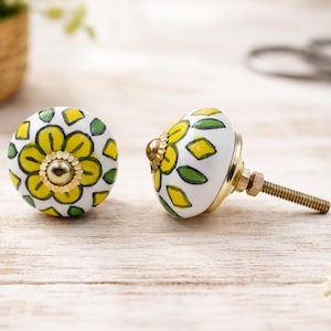 Op de afbeelding: Twee witte keramische kastknoppen met een bloemenontwerp. De knoppen hebben gele bloemen, groene bladeren en goudkleurige hardware. De knoppen liggen op een lichtgekleurd houten oppervlak.