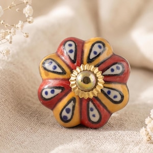 Könnte beinhalten: Ein blumenförmiger Keramik-Schrankknauf mit rotem, gelbem und blauem Design. Der Knauf hat eine goldfarbene Metallmitte und ist vor einem neutralen Hintergrund platziert. Die Blütenblätter haben eine Tropfenform mit blauen Punkten.