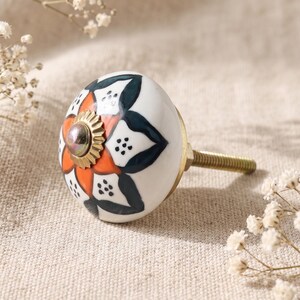 May include: A decorative cabinet knob with a white ceramic base featuring a floral design in orange and teal. The knob has a brass-colored metal base and screw, and a small, round, silver-colored center.