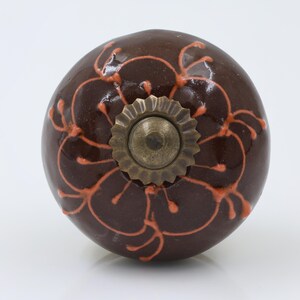 May include: A round, dark brown ceramic cabinet knob featuring a glossy finish and a raised, hand-painted orange floral pattern. The center of the knob is adorned with a scalloped, antique brass-toned metal hardware piece with a central screw head.