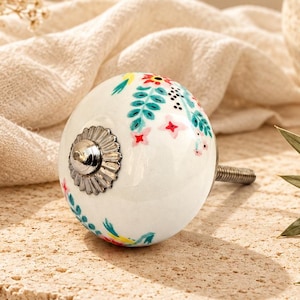 Op de afbeelding: Een ronde witte keramische meubelknop met een kleurrijk bloemenpatroon van groene bladeren, rode bloemen en kleine blauwe accenten. De knop heeft een zilverkleurige metalen voet met een geribbelde rand en een metalen schroef aan de achterkant.