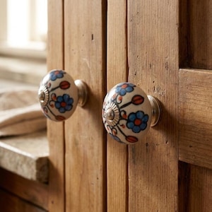 Könnte beinhalten: Zwei dekorative Schrankknöpfe mit floralem Design. Die Knöpfe sind weiß mit blauen und roten Blumen und silberfarbener Hardware. Sie sind an einer Holzschranktür befestigt.