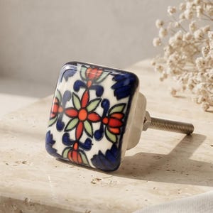 May include: A square ceramic cabinet knob featuring a hand-painted floral pattern in shades of deep blue, vibrant orange, and soft green. The knob has a smooth, glossy finish and is attached to a silver-toned metal bolt for secure installation.