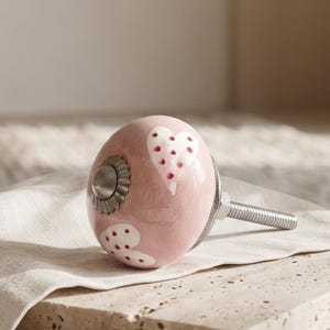 May include: A round, light pink ceramic cabinet knob featuring a pattern of white hearts with small red dots. The knob has a metallic silver-toned centre insert and a threaded metal bolt extending from the back for installation purposes.