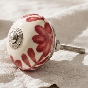 Può includere: Un pomello rotondo in ceramica per mobili con base color crema e motivi floreali e di foglie dipinti a mano in rosso scuro. Il pomello presenta un dettaglio centrale in metallo argentato e un perno filettato in metallo sul retro.