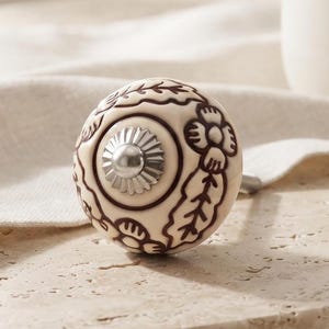 May include: A round ceramic cabinet knob featuring a cream-coloured base with dark brown floral and leaf patterns. The centre of the knob has a small, circular, silver-toned metal accent with a ridged, sunburst-like design and a smooth, rounded top.