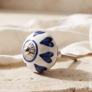 Peut inclure: Un bouton de meuble rond en céramique avec une base blanche ornée d'un motif peint à la main de cœurs bleu foncé rayonnant depuis un capuchon central en métal argenté. Le bouton comprend une tige filetée métallique à l'arrière.