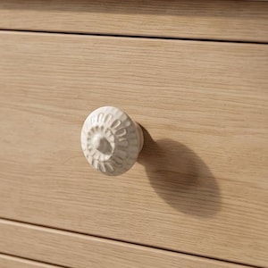 Puede incluir: Un pomo de armario redondo de cerámica blanca con un patrón floral radial texturizado, fijado a un cajón de madera clara. El pomo presenta un punto central elevado rodeado de ranuras talladas, aportando un detalle decorativo al mueble.