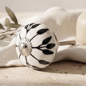 Può includere: Un pomello rotondo in ceramica per mobili con base bianca, un motivo botanico a foglie dipinto a mano in nero e un dettaglio centrale in metallo argentato. Il pomello include un perno metallico filettato sul retro per il fissaggio.