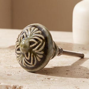 May include: A round ceramic cabinet knob featuring a glossy, mottled sage green glaze with a raised, cream-colored floral relief pattern on the front. The knob includes a silver-toned metal bolt extending from the back for installation purposes.