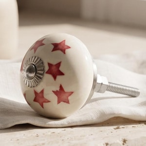 May include: A round, off-white ceramic cabinet knob featuring a pattern of small, reddish-pink stars. The knob has a central silver-toned metal hardware detail and a threaded metal bolt extending from the back for installation purposes.