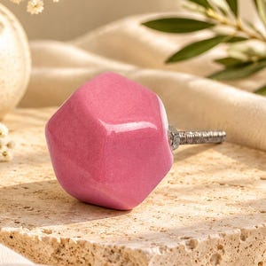 May include: A faceted, glossy pink ceramic cabinet knob with a metallic silver screw attachment. The knob has a geometric, multi-sided shape and a smooth, reflective surface, designed for installation on furniture or cabinetry doors.