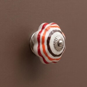 Könnte beinhalten: Ein runder, geriffelter Keramik-Schubladenknopf mit konzentrischen Ringen in Weiß, Orange und Dunkelbraun. Die Mitte des Knopfes ist mit einer kleinen, silberfarbenen Metallkappe mit geriffeltem Rand verziert, montiert auf einer matten, braunen Front.