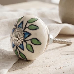 May include: A round ceramic cabinet knob featuring a hand-painted floral pattern with blue petals and green leaves on an off-white background. The centre of the knob has a silver-toned metal accent, and a threaded metal bolt extends from the back.