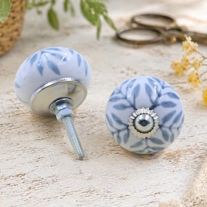 Puede incluir: Dos pomos de cerámica para armarios con base blanca y un patrón de hojas azules. Cada pomo tiene una base de metal plateado y un perno roscado para la instalación. Los pomos son redondos y tienen un diseño floral decorativo.