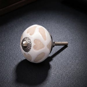 May include: A white ceramic cabinet knob with a beige heart pattern. The knob features a silver-coloured metal base and screw. Suitable for drawers, cabinets, or furniture. The knob is approximately 3.8 cm in diameter.