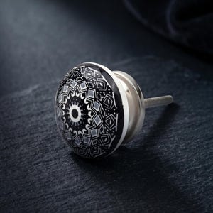 May include: A black and white patterned cabinet knob with a silver metal base. The knob's design features a central circular motif surrounded by geometric shapes. The knob is designed for easy installation on furniture.