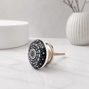 Puede incluir: Pomo de armario de cerámica en blanco y negro con un diseño de mandala decorativo. El pomo tiene una base y un tornillo de metal plateado. El pomo redondo tiene un acabado brillante. El fondo es gris claro.