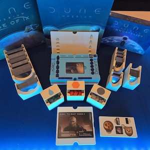 Puede incluir: Un tablero de juego con un diseño azul y blanco, etiquetado como "Deck" y "Discard". El tablero está rodeado de contenedores de plástico blanco con varias piezas de juego, incluyendo piezas naranjas, grises y azules. El juego se llama "Dune: Imperium Rise of Ix".