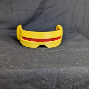 May include: A yellow visor with a red mesh detail. The visor has a curved shape and is designed to fit over the eyes. The item is made of a solid material and is likely a costume accessory.