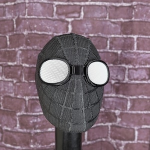 May include: A black Spider-Man mask with white-lensed goggles. The mask has a textured, web-like pattern and is displayed on a black stand against a brick wall background. The goggles are round and have a perforated design.
