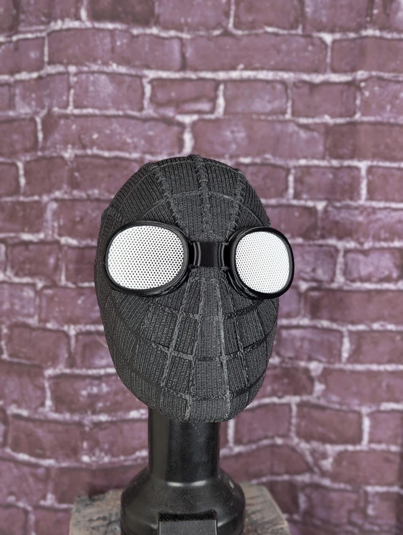 May include: A black Spider-Man mask with white-lensed goggles. The mask has a textured, web-like pattern and is displayed on a black stand against a brick wall background. The goggles are round and have a perforated design.