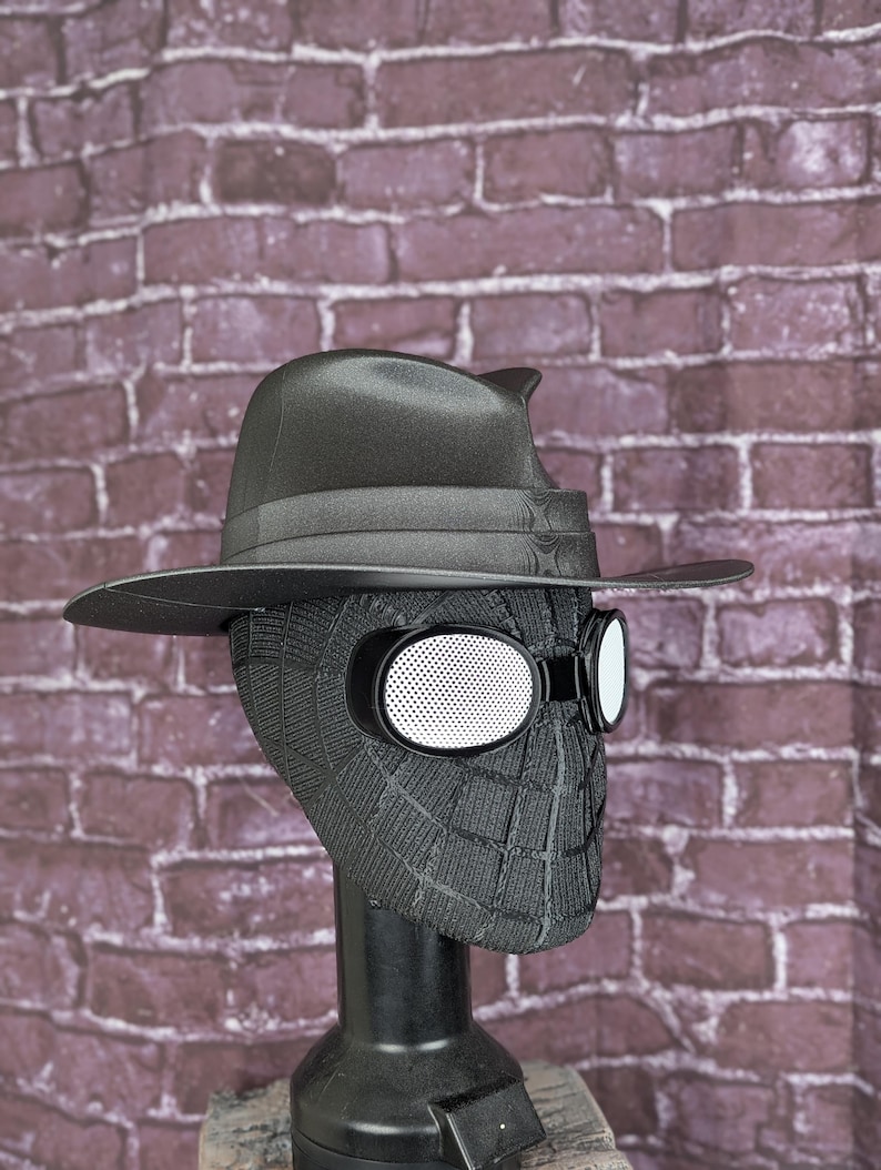 May include: A black Spider-Man mask with a fedora hat and white-rimmed goggles. The mask has a webbed design and is displayed on a black stand. The hat has a curved brim and a ribbon around the crown.