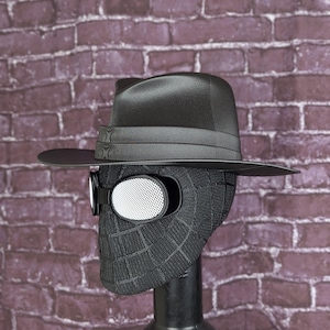 May include: A black fedora hat rests atop a black Spider-Man mask with white mesh eye lenses. The mask is on a black mannequin head, set against a brick wall background. The hat has a black band and a wide brim.