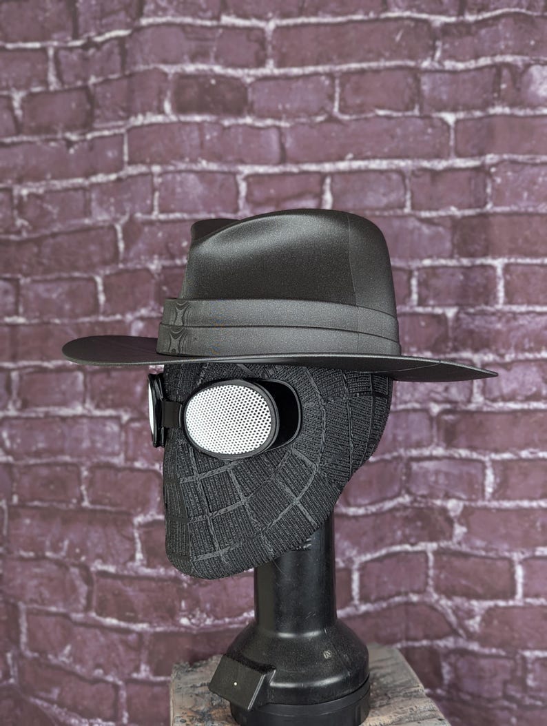 May include: A black fedora hat rests atop a black Spider-Man mask with white mesh eye lenses. The mask is on a black mannequin head, set against a brick wall background. The hat has a black band and a wide brim.