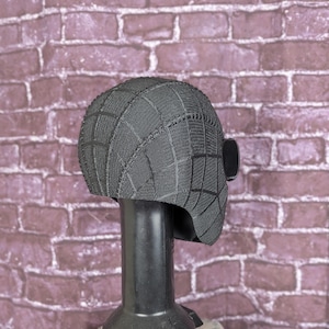 May include: A black, textured mask with a web pattern, displayed on a black stand. The mask has a rounded shape with a rectangular eye piece on the side. The background is a brick wall.