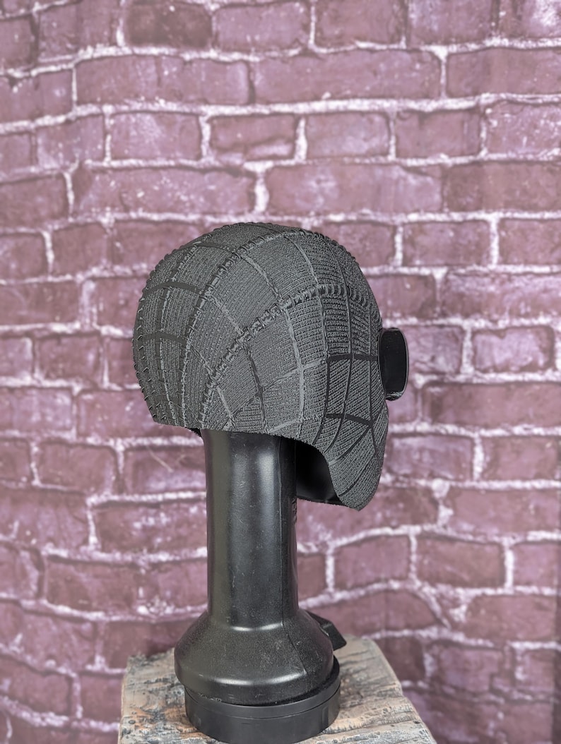 May include: A black, textured mask with a web pattern, displayed on a black stand. The mask has a rounded shape with a rectangular eye piece on the side. The background is a brick wall.