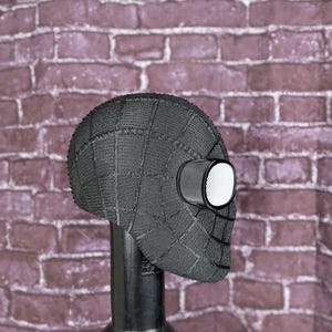May include: A black Spider-Man mask with a textured, web-like pattern. The mask features large, white-rimmed eye lenses and is displayed on a black stand. The background is a brick wall.