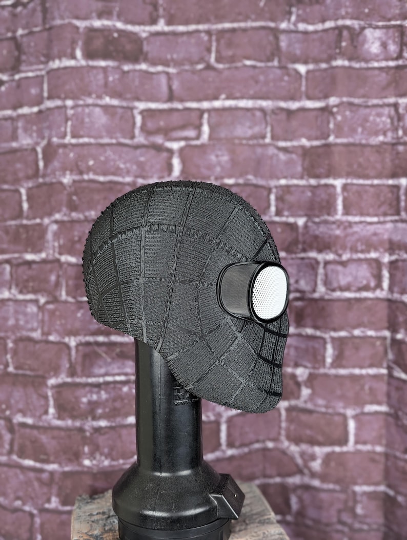 May include: A black Spider-Man mask with a textured, web-like pattern. The mask features large, white-rimmed eye lenses and is displayed on a black stand. The background is a brick wall.