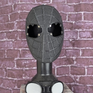 May include: A black Spider-Man mask with a textured web pattern, displayed on a black stand. The mask has large eye openings and is paired with white-rimmed goggles with a mesh design. The background is a brick wall.