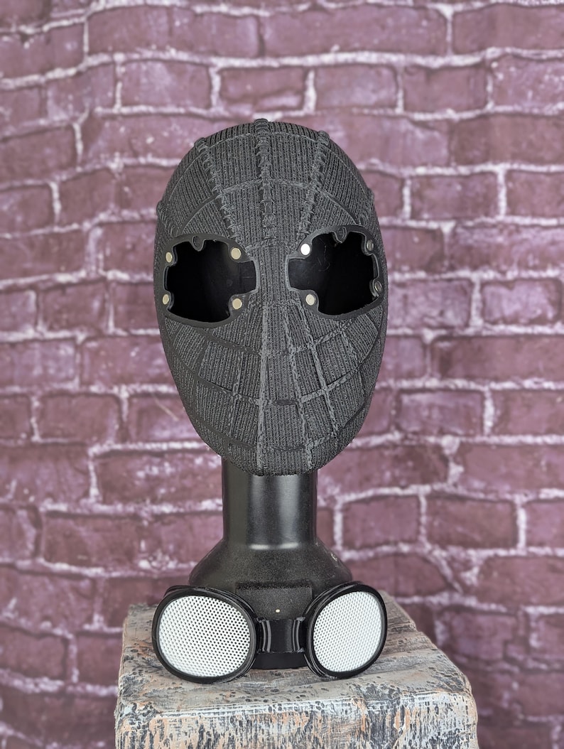 May include: A black Spider-Man mask with a textured web pattern, displayed on a black stand. The mask has large eye openings and is paired with white-rimmed goggles with a mesh design. The background is a brick wall.