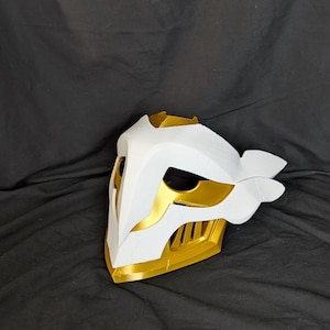 May include: A white and gold mask with a stylized design. The mask features a white base with gold accents on the edges, eye openings, and a central crest. The mask has a smooth, clean finish and appears to be 3D-printed.