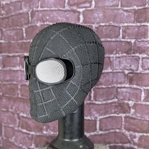 May include: A black, textured mask with a grid pattern, featuring white mesh eye lenses and a black strap. The mask is displayed on a black stand against a brick wall background. This item is likely a costume or cosplay accessory.