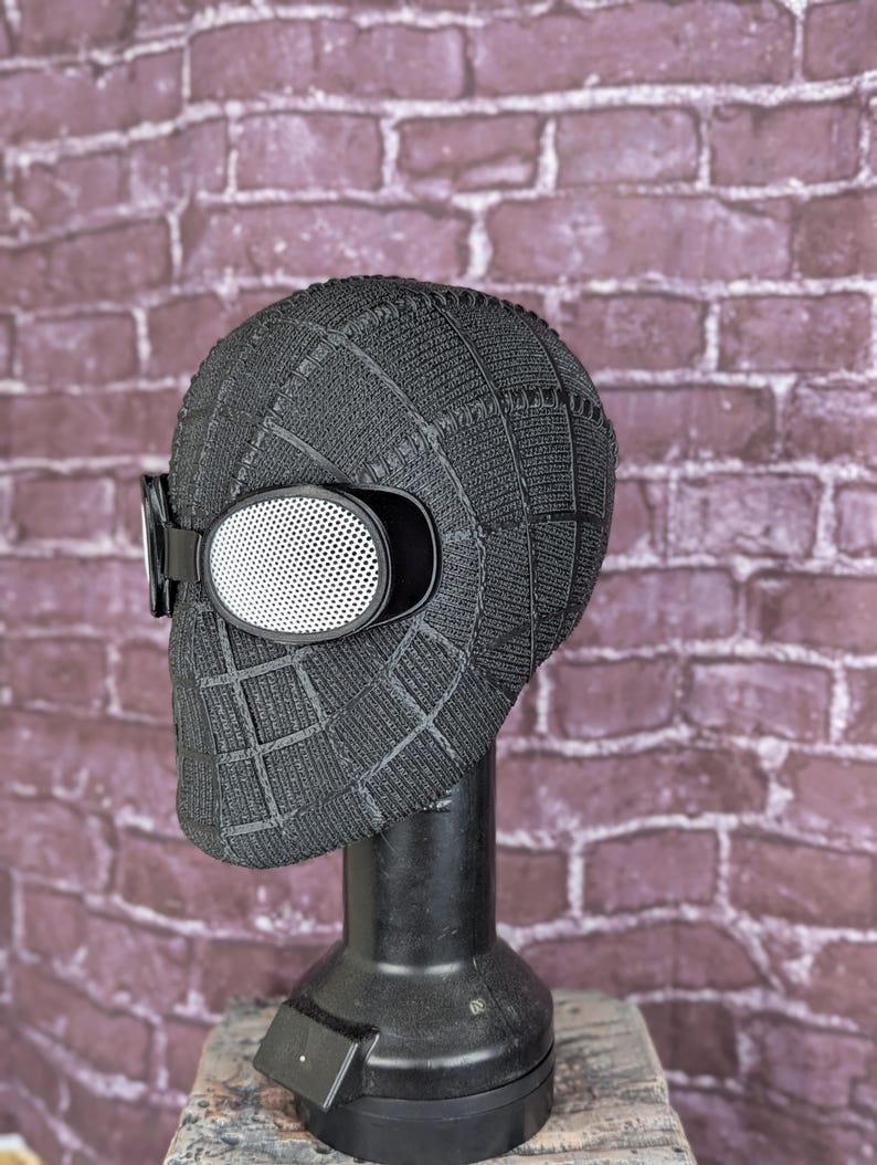 May include: A black, textured mask with a grid pattern, featuring white mesh eye lenses and a black strap. The mask is displayed on a black stand against a brick wall background. This item is likely a costume or cosplay accessory.